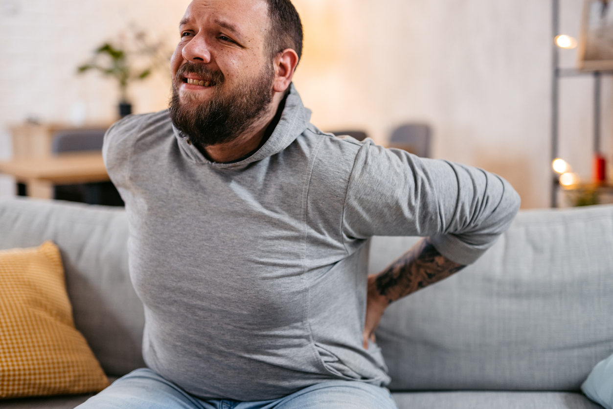 Person with pained expression sitting on home sofa with both hands on lower back